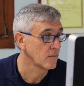 Un uomo di mezza età con capelli grigi e occhiali blu sta guardando lo schermo di un computer in un ufficio al chiuso.