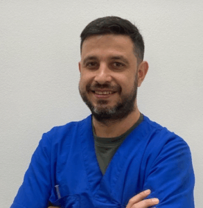 Un uomo con capelli corti e barba scura sorride e sta in piedi con le braccia incrociate. Indossa un camice medico blu e si trova di fronte a una parete bianca.