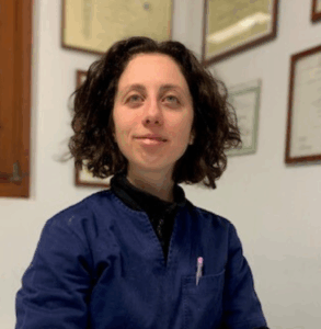 Una persona con i capelli ricci castani che indossa una giacca blu navy siede in casa, sorridendo leggermente. Sulla parete alle sue spalle sono esposti certificati o diplomi incorniciati.