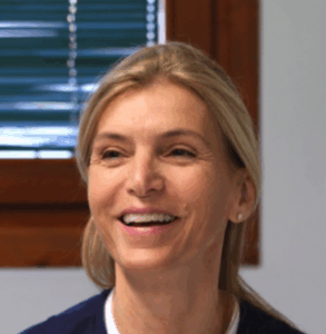 Una donna con i capelli biondi sorride mentre è seduta in casa vicino a una finestra con tende verdi e telaio in legno. Indossa un top scuro e orecchini a perno.