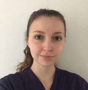 Una giovane donna con lunghi capelli castani legati all'indietro, che indossa un top scuro, è in piedi di fronte a un semplice muro bianco e sorride dolcemente alla telecamera.