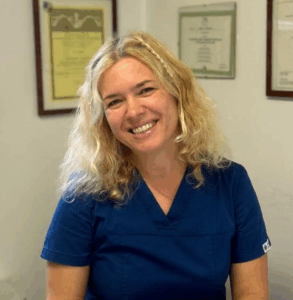 Una donna dai capelli ricci e biondi e dal sorriso smagliante indossa un camice medico blu e siede in casa con certificati incorniciati sulla parete alle sue spalle.