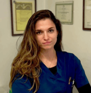 Una donna dai lunghi capelli castani che indossa un camice medico blu siede in un ufficio con certificati incorniciati sulla parete alle sue spalle. È rivolta verso la telecamera con un'espressione neutra.