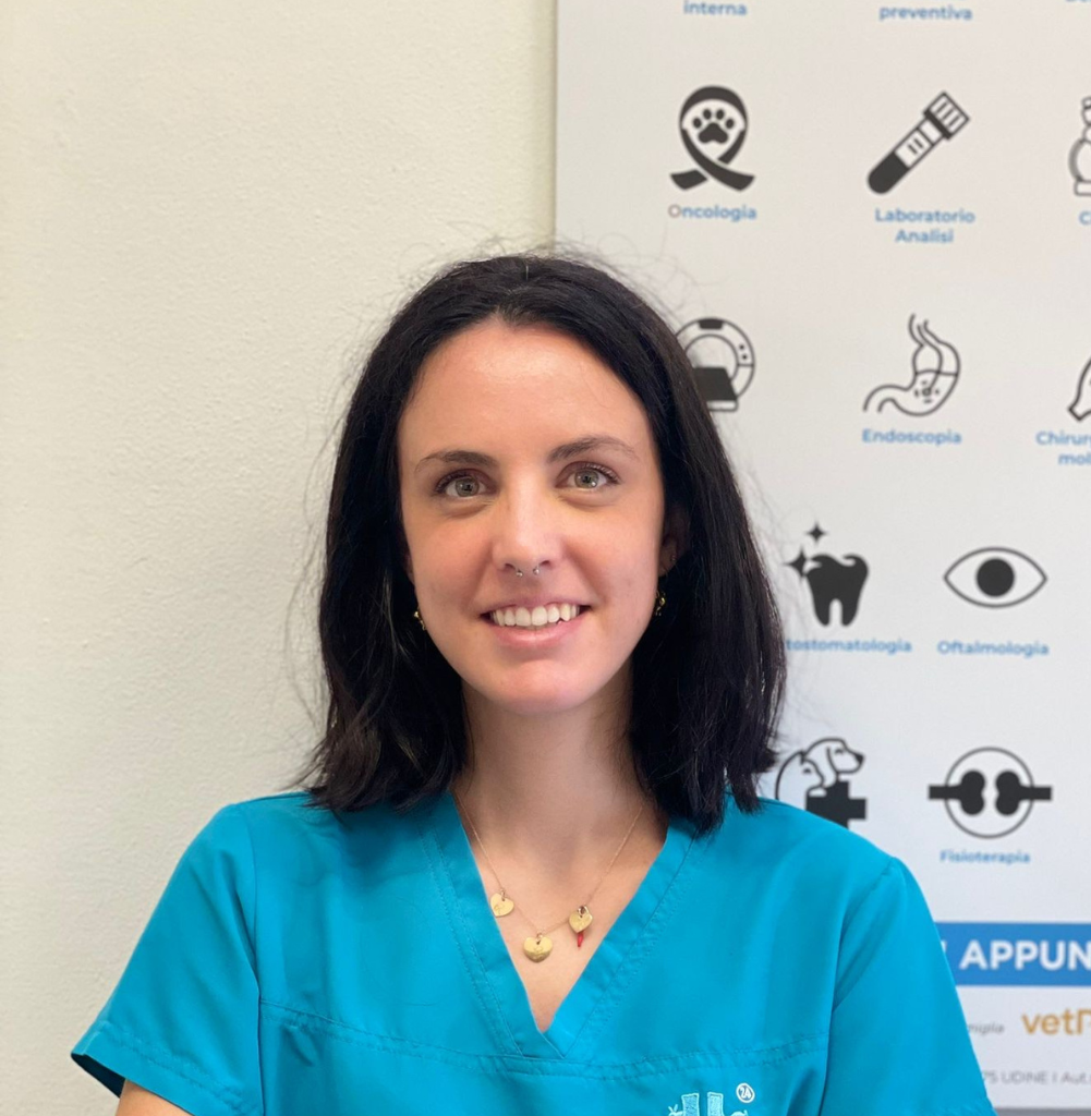 Una donna con i capelli scuri lunghi fino alle spalle, che indossa un camice medico blu e una collana d'oro, sorride alla telecamera. Dietro di lei c'è una parete bianca e un poster con icone e testi medici.