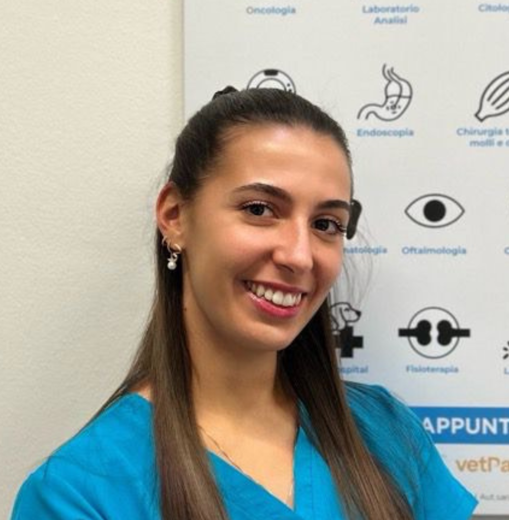 Una donna in camice blu sorride alla telecamera davanti a una parete con icone e testi medici.