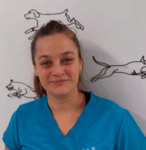 Una donna in camice blu sorride alla telecamera, in piedi davanti a una parete bianca decorata con disegni a linee nere di cani in corsa.